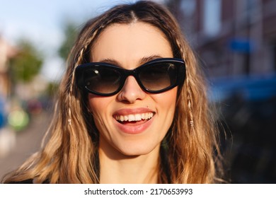 Pensive Blonde Woman In Black Glasses Posing On Blur City Street Background. Outdoor Shot Of Happy Hippie Lady With Two Thin Braids And Wave Hair. Coachella Or Boho Freedom Style.