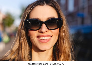 Pensive Blonde Woman In Black Glasses Posing On Blur City Street Background. Outdoor Shot Of Happy Hippie Lady With Two Thin Braids And Wave Hair. Coachella Or Boho Freedom Style.