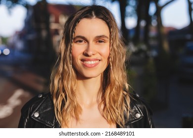 Pensive blonde woman in black leather jacket posing on blur city street background. Outdoor shot of happy hippie lady with two thin braids and wave hair. Coachella or boho freedom style.