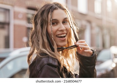 Pensive blonde woman in black leather jacket put off black glasses and turn around posing on street background. Outdoor shot of happy hippie lady with two thin braids and wave hair. Boho freedom style