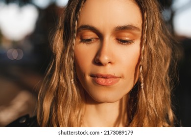 Pensive Blonde Woman In Black Leather Jacket Posing On Blur City Street Background. Outdoor Shot Of Happy Hippie Lady With Two Thin Braids And Wave Hair. Coachella Or Boho Freedom Style. Look At Down.