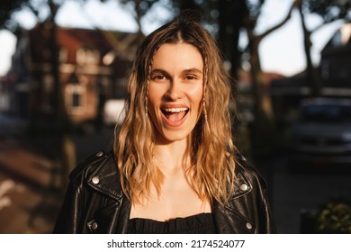 Pensive Blonde Woman In Black Leather Jacket And Dress With Open Mouth Posing On Street. Outdoor Shot Of Happy Hippie Lady With Two Thin Braids And Wave Hair. Coachella Or Boho Freedom Style.