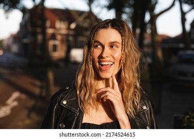 Pensive Blonde Woman In Black Leather Jacket And Dress With Open Mouth And Finger Near Face, Good Idea. Outdoor Shot Of Happy Hippie Lady With Two Thin Braids And Wave Hair. Coachella Freedom Style.