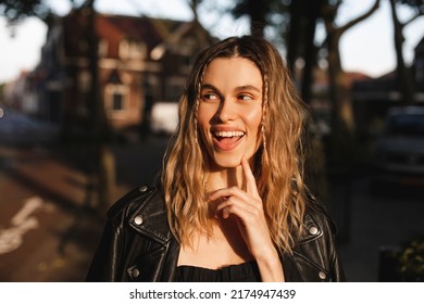 Pensive Blonde Woman In Black Leather Jacket And Dress With Open Mouth And Finger Near Face, Good Idea. Outdoor Shot Of Happy Hippie Lady With Two Thin Braids And Wave Hair. Coachella Freedom Style.