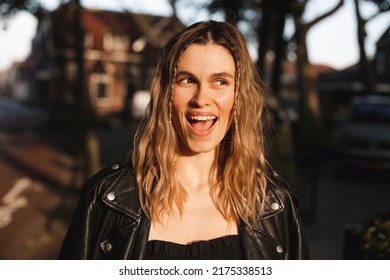 Pensive Blonde Woman In Black Leather Jacket And Dress With Open Mouth Posing On Street. Outdoor Shot Of Happy Hippie Lady With Two Thin Braids And Wave Hair. Coachella Or Boho Freedom Style.