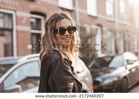 Pensive blonde woman in black leather jacket, black glasses and turn back posing on street background. Outdoor shot of happy hippie lady with two thin braids and wave hair. Boho freedom style