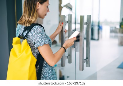 Pensive blonde woman in casual wear with backpack open door using smartphone with mock up screen application, serious female unlocking security system via mobile phone checking code for entrance