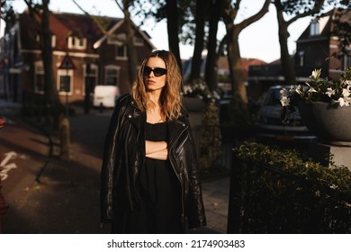 Pensive Blonde Woman Wear Black Dress, Glasses, Leather Jacket And Touch It, Posing. Outdoor Shot Of Calm Sensual Hippie Lady With Two Thin Braids And Wave Hair. Coachella Or Boho Freedom Style.