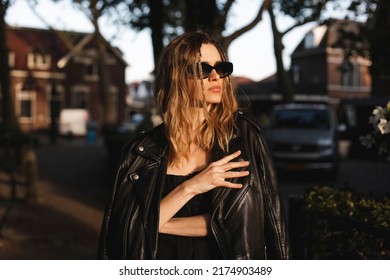 Pensive Blonde Woman Wear Black Dress, Glasses, Leather Jacket And Touch It, Posing. Outdoor Shot Of Calm Sensual Hippie Lady With Two Thin Braids And Wave Hair. Coachella Or Boho Freedom Style.