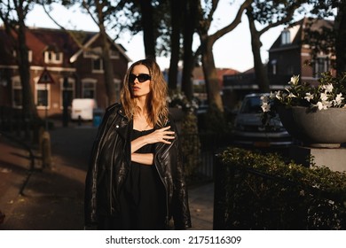 Pensive Blonde Woman Wear Black Dress, Glasses, Leather Jacket And Touch It, Posing. Outdoor Shot Of Calm Sensual Hippie Lady With Two Thin Braids And Wave Hair. Coachella Or Boho Freedom Style.