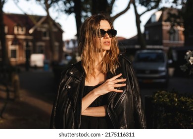 Pensive Blonde Woman Wear Black Dress, Glasses, Leather Jacket And Touch It, Posing. Outdoor Shot Of Calm Sensual Hippie Lady With Two Thin Braids And Wave Hair. Coachella Or Boho Freedom Style.