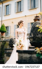 Pensive bride in a pink dress with a bouquet of flowers stands with her head bowed on the steps of an ancient villa. Lake Como, Italy