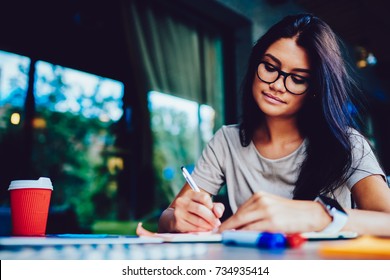 Pensive Brunette Female Student In Eyeglasses Concentrated On Homework Writing Essay In Cafe Interior, Talented Designer Drawing Sketch And Graphic For Project Having Creative Idea Using Stationery