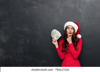 Pensive brunette woman in red blouse and christmas hat holding money and looking up over black background