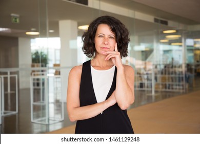 Pensive Business Woman With Folded Arms Posing In Office Hall. Thoughtful Businesswoman In Casual Standing Near Glass Wall, Touching Chin And Looking At Camera. Business Portrait Concept