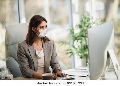 A Pensive Business Woman With Surgical Mask Sitting Alone In Her Office And Working On Computer.