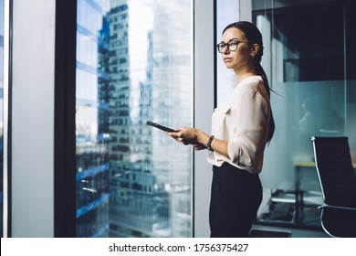 Pensive businesswoman dressed in formal apparel standing near panoramic window in enterprise company and thinking about trade investment,thoughtful female entrepreneur in eyewear for vision protection