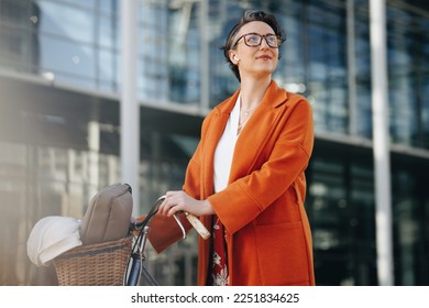 Pensive businesswoman pushing her bike during her morning commute in the city. Mature business woman looking away thoughtfully while walking to her office with a bicycle.