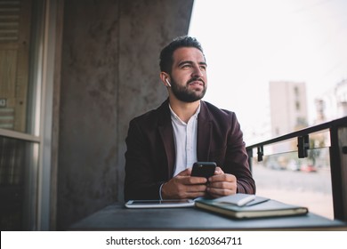 Pensive caucasian male entrepreneur in earphones enjoying playlist songs via smartphone and bluetooth connected accessory, confident businessman using smartphone for chatting and listening audiobook - Powered by Shutterstock