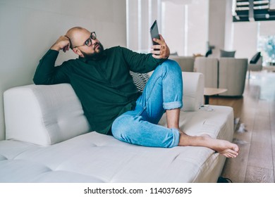 Pensive Caucasian Male In Spectacles Sitting On Cozy Sofa At Partment Watching Video On Portable Pc, Hipster Guy In Eyewear Reading Ebook Via Digital Tablet Using Wireless Connection At Home