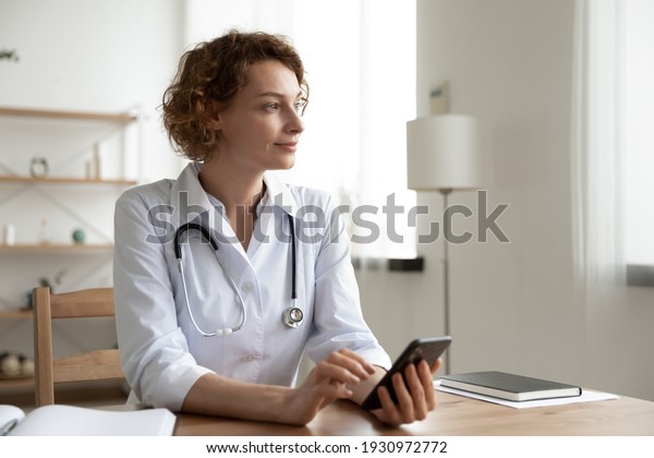 Pensive Caucasian woman nurse sit at desk in\
hospital use cellphone look in distance dreaming visualizing.\
Dreamy female doctor consult patient online on smartphone. Medicine\
and technology concept.