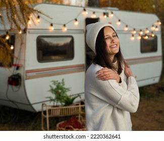 A pensive Caucasian woman in a white knitted sweater and a hat is resting near a motorhome in a warm autumn. Travel by camper.
