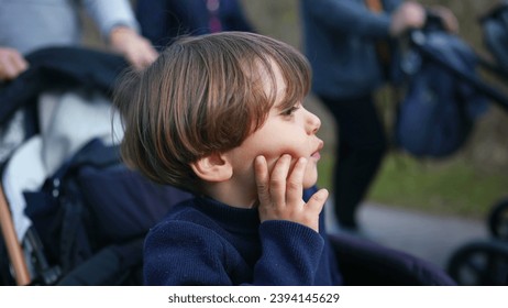 Pensive child thinking while seated on stroller with hand in chin. Young boy observing world with thoughtful expression while being carried in carriage