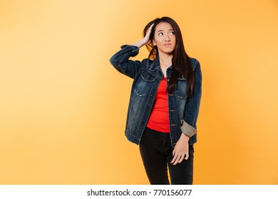 Pensive confused asian woman in denim jacket holding head and looking away over yellow background - Shutterstock ID 770156077