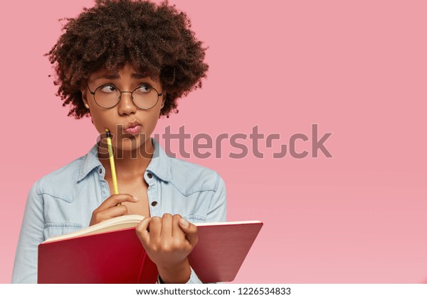 Pensive confused dark skinned woman purses lips in bewilderment, writes notes in notepad, thinks on creating new article for personal blog, wears round transparent glasses poses indoor with copy space