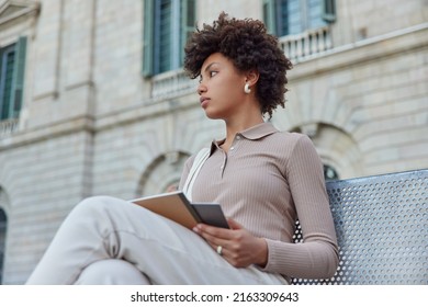 Pensive Curly Haired Woman Poses On Bench With Notebooks Thinks On Writing Essay On Topic Prepares For Exams Wears Casual Clothes Focused Into Distance Poses Outdoors Near Old Histotic Building.