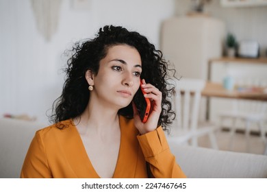 Pensive curly young woman in orange blouse talks by phone with perplexed face standing home against blurry kitchen. Sad caucasian housewife got bad news. Candid people emotions. Sorrow - Powered by Shutterstock