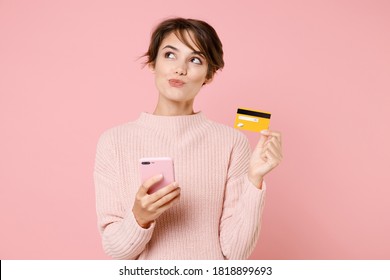 Pensive Cute Young Woman 20s In Knitted Casual Sweater Using Mobile Cell Phone, Hold Credit Bank Card Doing Online Shopping Isolated On Pastel Pink Background Studio Portrait. People Lifestyle Concept