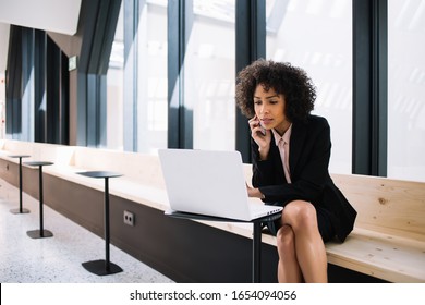 Pensive dark skinned businesswoman making booking via laptop computer talking with operator on mobile phone, confident african american female entrepreneur using technology for project creation