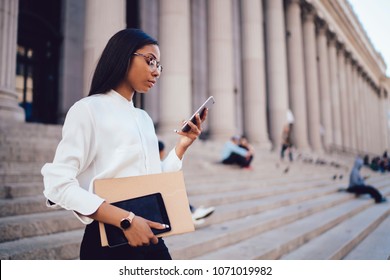 Pensive Dark Skinned Student Of Faculty Of Law Checking Mail And Reading Notification With Financial News On Smartphone Device Connected To Smartphone Standing Outdoors Near University Building