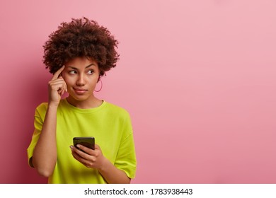 Pensive dark skinned woman keeps finger on temple, looks aside thoughtfully, uses new gadget for first time, dressed casually, tries to remember name of product before browsing internet, has idea