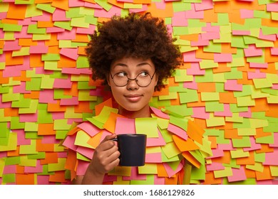 Pensive dark skinned woman works in office, has coffee break, holds cup of tea, looks aside, thinks about new project, wears round transparent glasses, sticks head through wall, adhesive notes around