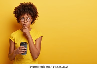 Pensive displeased woman with dark skin, holds chin and looks away, contemplates about something, wears yellow t shirt, holds disposable cup of coffee, smirks face and being upset by bad thoughts
