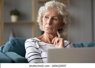 Pensive distracted old woman sitting on sofa with laptop look out the window feels puzzled about electronic device usage, thinking about retired life, waiting video call from grown up children concept