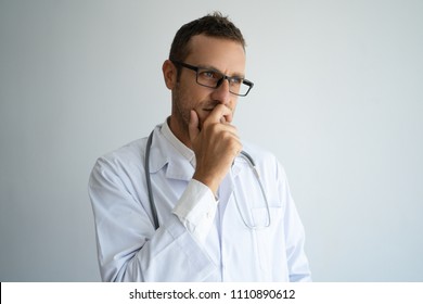 Pensive Doctor In Glasses Reflecting On Patient Treatment. Focused Practitioner Leaning Head On Hand, Covering Mouth With Fist. Decision Making And Medicine Concept