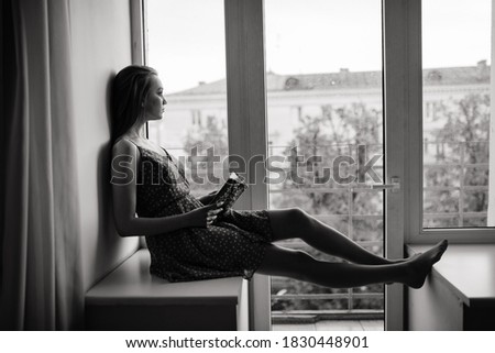 Pensive dreamy female holding book at home looking at big window dreaming, thoughtful young woman, student