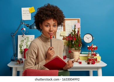 Pensive Dreamy Young Female Writes Down Notes, Creats Essay On Interesting Topic, Holds Pencil And Opened Red Notebook, Dressed Casually, Poses In Own Cabinet Decorated For Christmas Or New Year.