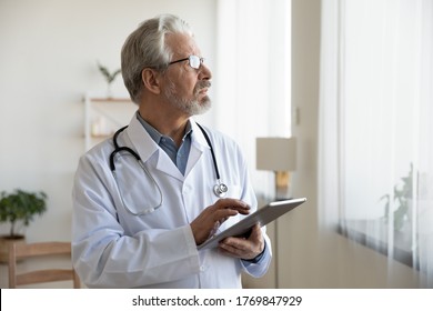 Pensive Elderly Male Doctor In White Medical Uniform Work On Modern Tablet Look In Distance Thinking Pondering, Thoughtful Mature Man GP Physician Use Pad Gadget Make Decision In Hospital