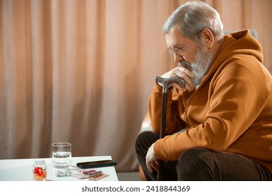 Pensive elderly man with cane sitting near a table with a glass of water, smartphone, and medications. Concept of health and medical care, aging, medicine, treatment - Powered by Shutterstock