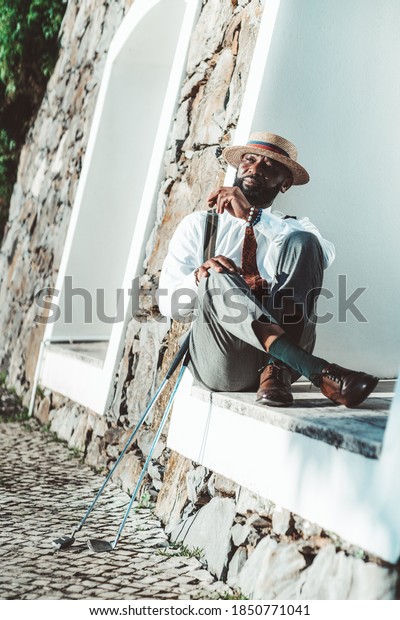 A pensive elegant black retrosexual guy in a\
vintage costume and a hat is having rest and sitting in a recess of\
a building with a cigar in his hand and two golf clubs near him on\
paving-stone