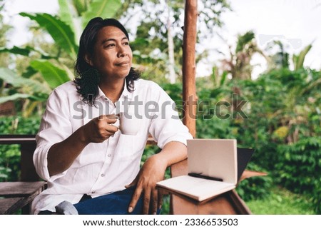 Pensive ethnic male entrepreneur with long dark hair in casual outfit enjoying own product coffee while sitting at counter with notepad on veranda and looking at trees growing on farm