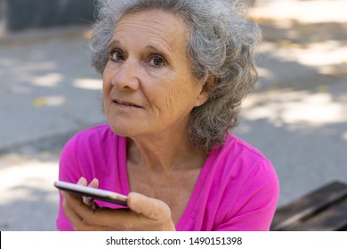 Pensive Excited Old Lady Using Speaker While Talking On Phone. Upset Senior Grey Haired Woman In Casual Sitting On Park Bench And Holding Smartphone Near Face. Voice Connection Concept