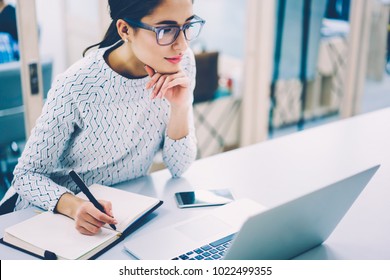 Pensive female administrative assistant checking report on laptop reading information and noting data,skilled woman watching online video online pondering on creating publication writing in notepad