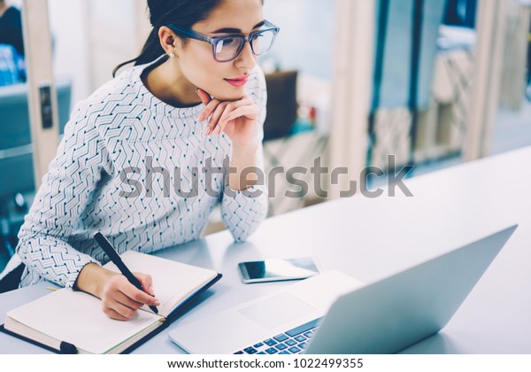 Pensive female administrative assistant checking\
report on laptop reading information and noting data,skilled woman\
watching online video online pondering on creating publication\
writing in notepad