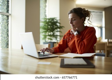 Pensive female typing text on keyboard of modern laptop computer in searching useful information for coursework.Smart student sitting in university with literature books and optical spectacles in hand