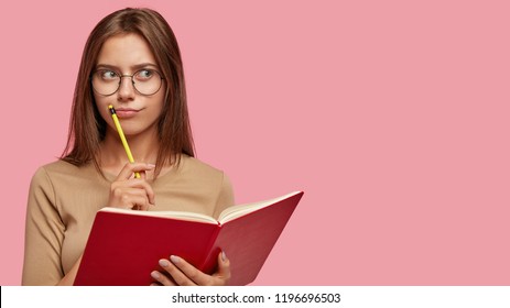 Pensive female writer ponders on new ideas, holds red notebook and pencil near mouth, has thoughtful expression, wears big round glasses, isolated over pink background with copy space on right - Powered by Shutterstock